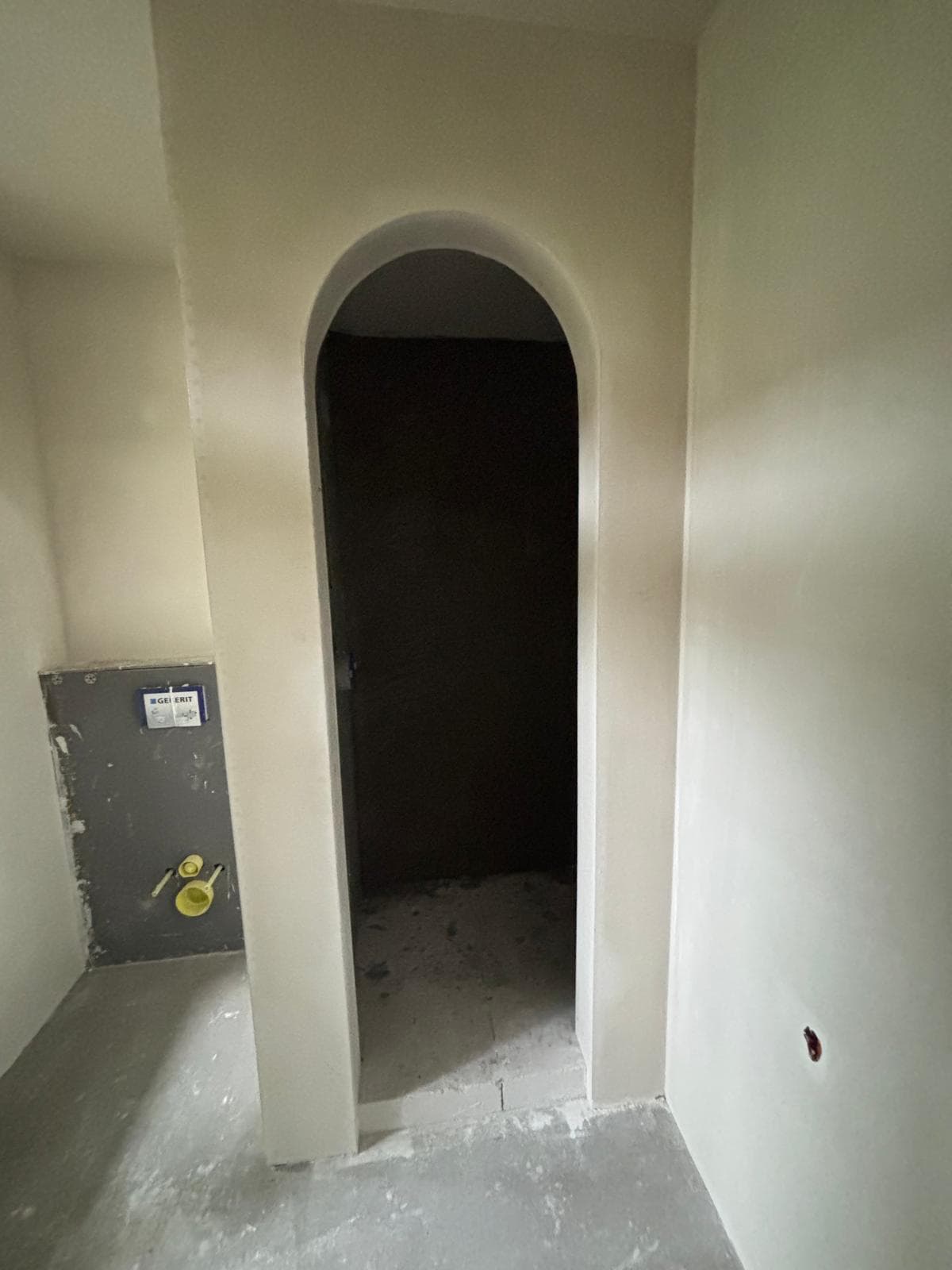 Arched doorway in an unfinished room with plastered walls and wall-hung toilet plumbing fixtures.