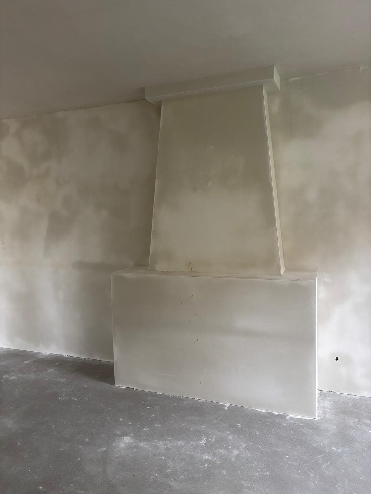 A minimalist white plastered fireplace surround in an unfinished room with a concrete floor.